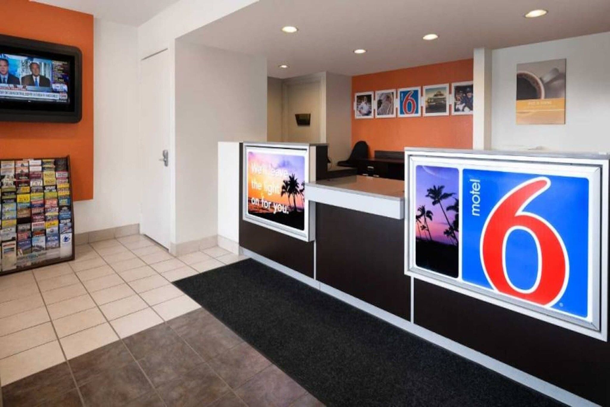 Área de recepção de hotel com tela digital exibindo um canal de notícias, balcão de atendimento e parede com fotografias emolduradas.