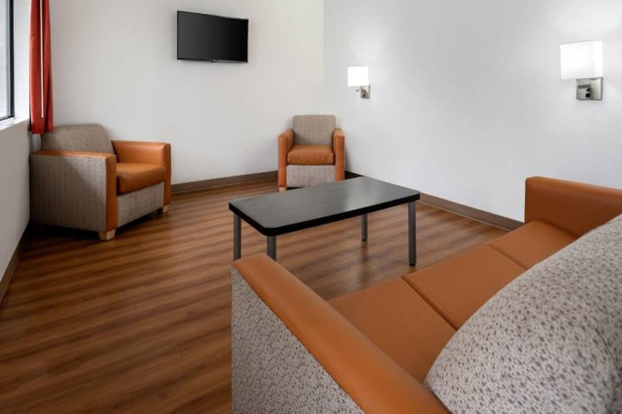 A modern hotel room with a wooden floor, two sofas, a coffee table, and a flat-screen TV mounted on the wall.