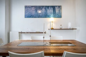 Un comedor con una mesa de madera, sillas blancas, una mesa con cubierta de vidrio, un cuadro colgado en la pared, una lámpara de pie y una estantería con adornos.