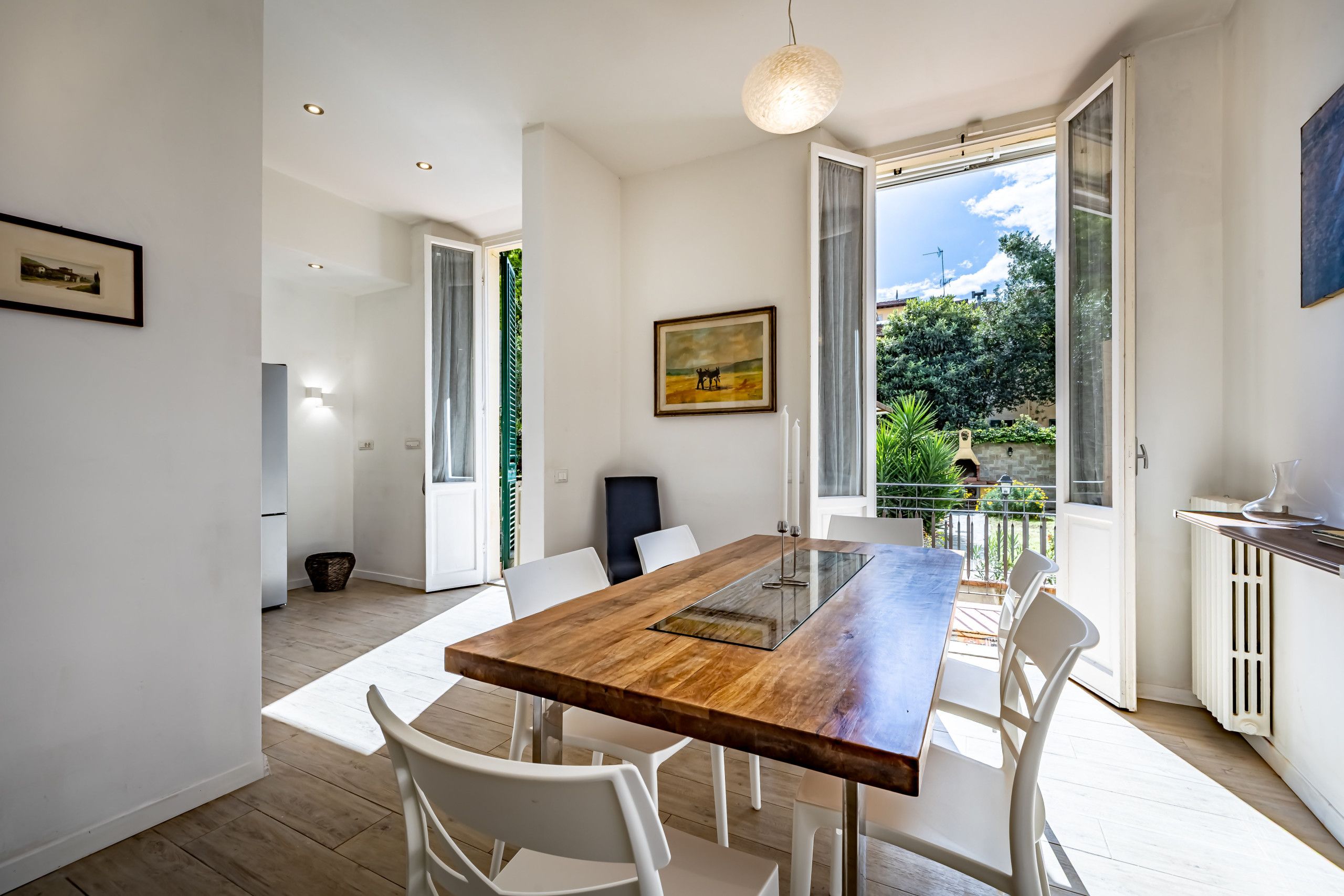 A modern dining area with a wooden table and white chairs, a painting on the wall, and a view of greenery outside.