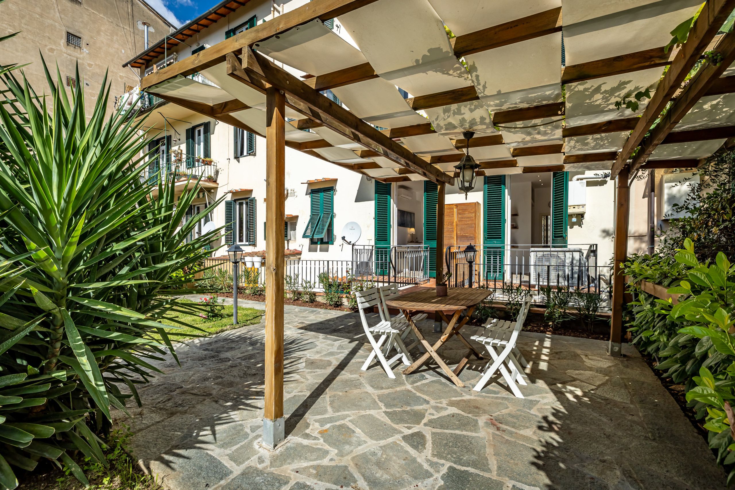 Area patio esterna con pergola in legno, pavimento in pietra e mobili bianchi.