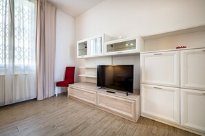 Een moderne woonkamer met een flatscreen televisie op een houten kast, een rode fauteuil en een groot raam met dunne gordijnen.
