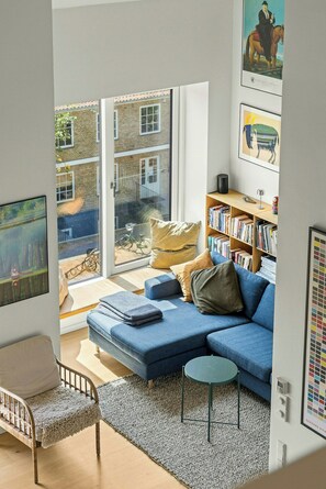 Ein modernes Wohnzimmer mit einem blauen Sofa, einem kleinen runden Tisch, einem Bücherregal und einem großen Fenster mit Blick auf Gebäude.