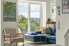 A modern living room with a blue sofa, a wicker chair, a small side table, and a bookshelf filled with books. - Østerbro flat with views over Copenhagen (København)