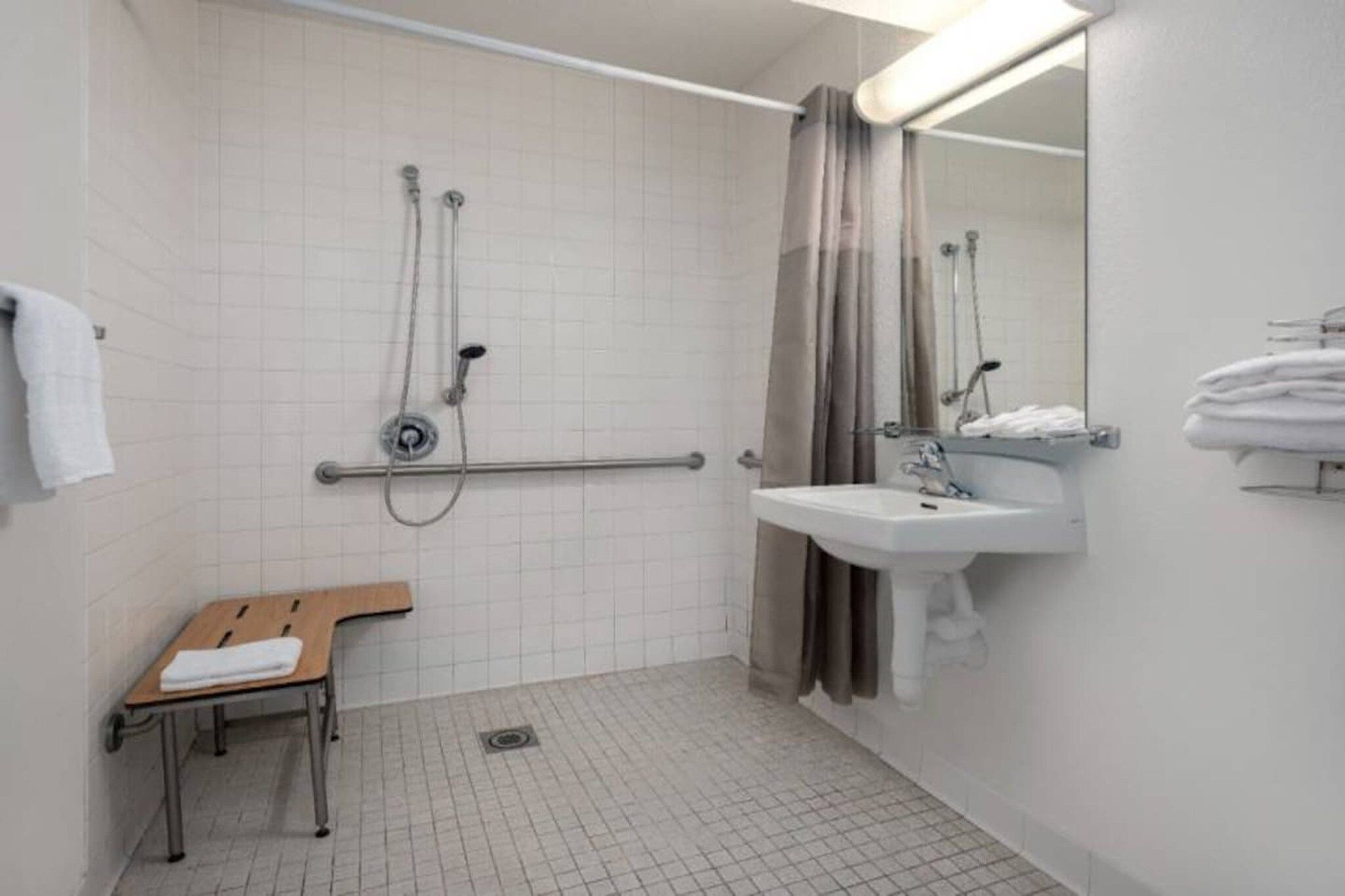 A bathroom with a shower, a sink, a mirror, a towel rack, and a bench.