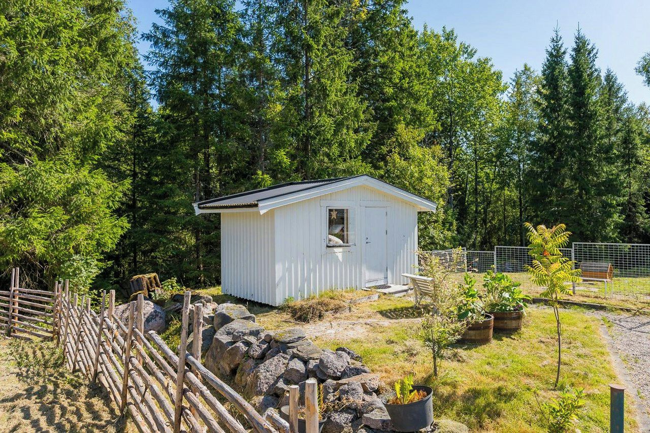 Pieni valkoinen puinen rakennus, jossa on ovi ja ikkunat. Sen ympärillä on puinen aita ja kivimuuri, ja se sijaitsee metsäisellä alueella.