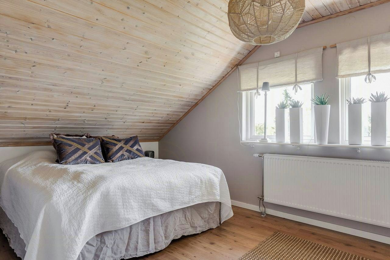 Ett sovrum med ett sluttande tak, en säng med ett vitt överkast, två kuddar, en vävd lampa, ett fönster med persienner och krukväxter.