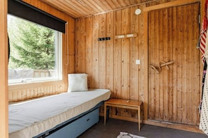 A wooden cabin room with a bed, a bench, a window with a view of trees, and a door with a handle.