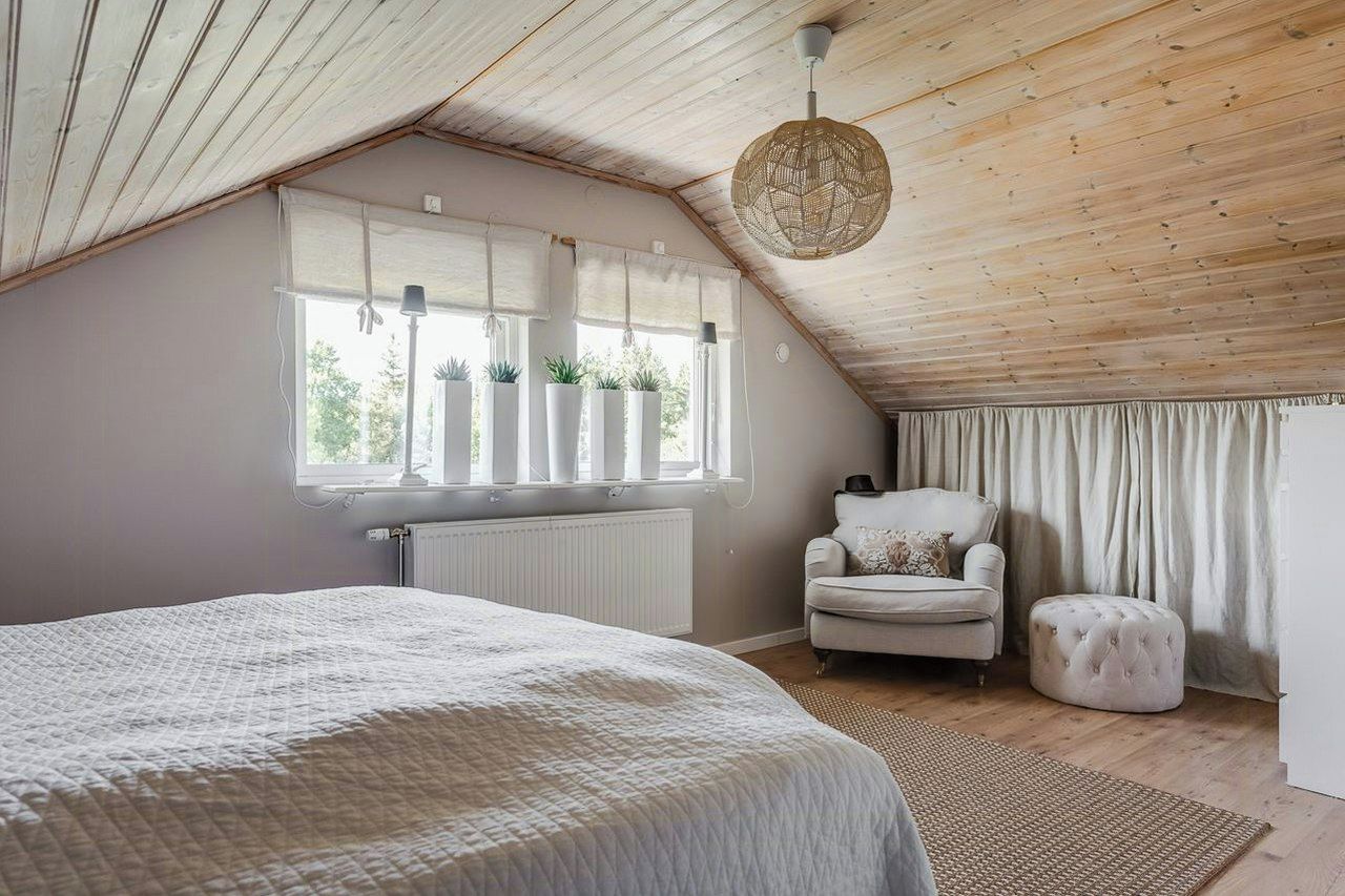 A bedroom with a sloped ceiling, a bed with a white quilt, a chair, a round ottoman, and a window with plants.