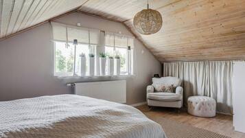 A bedroom with a sloped ceiling, a bed with a white quilt, a chair, a round ottoman, and a window with plants.