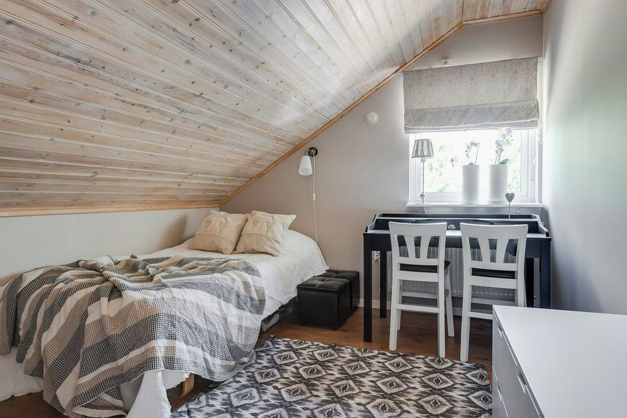 Un dormitorio con techo inclinado, una cama con manta estampada, un escritorio con sillas y una ventana con vista a follaje.