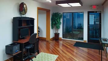 Una habitación con escritorio, silla, computadora, estantería, planta en maceta y vistas a través de un ventanal.