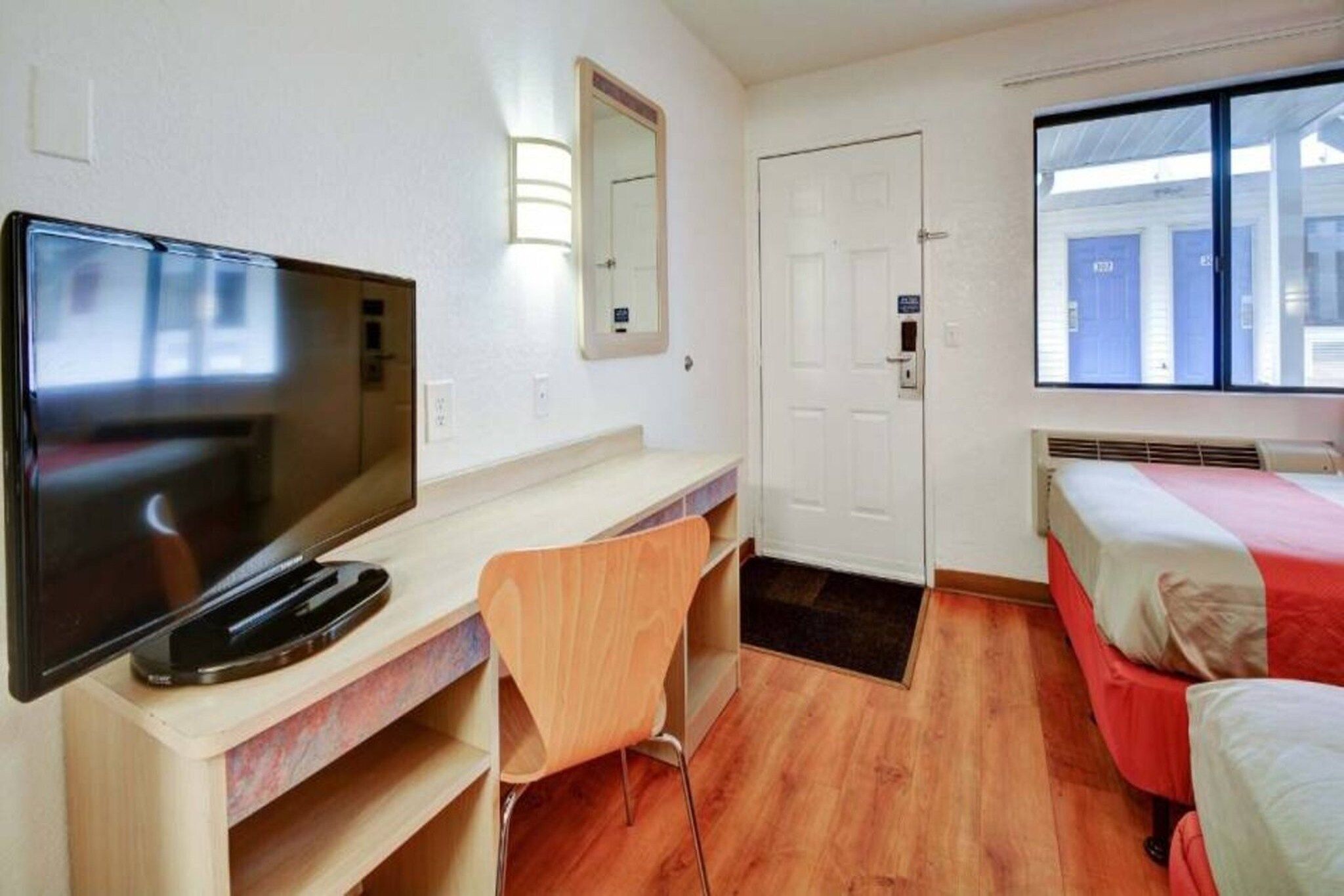 A hotel room with a television, a desk, a chair, a bed, a door, and a window.