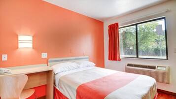 Habitación de hotel con cama, escritorio, silla, ventana con vistas a árboles y lámpara de pared.