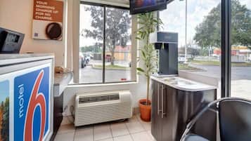 A motel interior with a reception desk, a television mounted on the wall, a window with a view of the parking lot, and a potted plant.