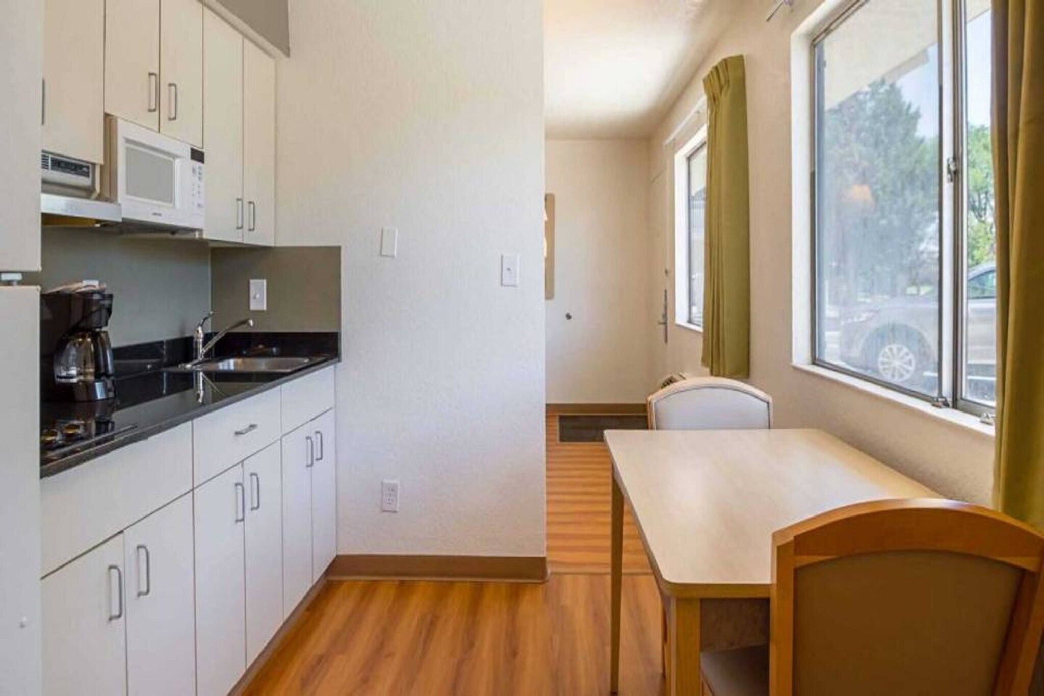 Cocina compacta con microondas, cafetera y un lavamanos pequeño. Hay una zona de comedor con mesa y sillas, y una ventana con cortinas.