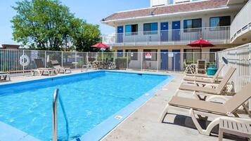 Piscina con sillas de descanso y sombrillas, rodeada por una zona vallada y un edificio al fondo.