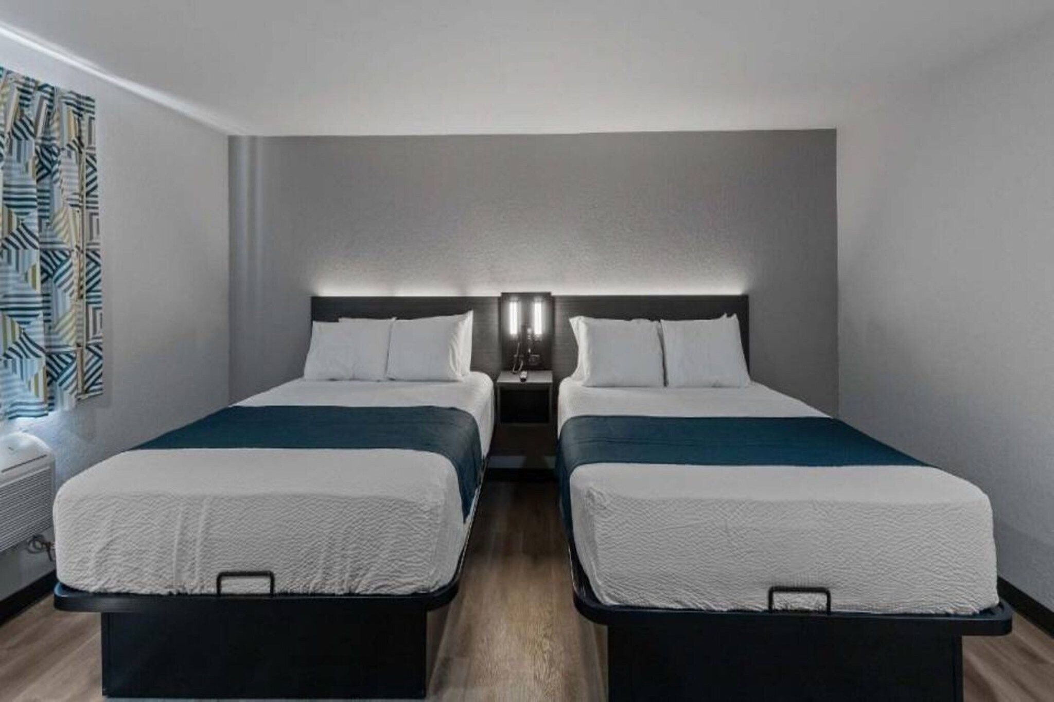 Ein Hotelzimmer mit zwei Betten, einem Nachttisch mit Lampe und einem Fenster mit Vorhängen.