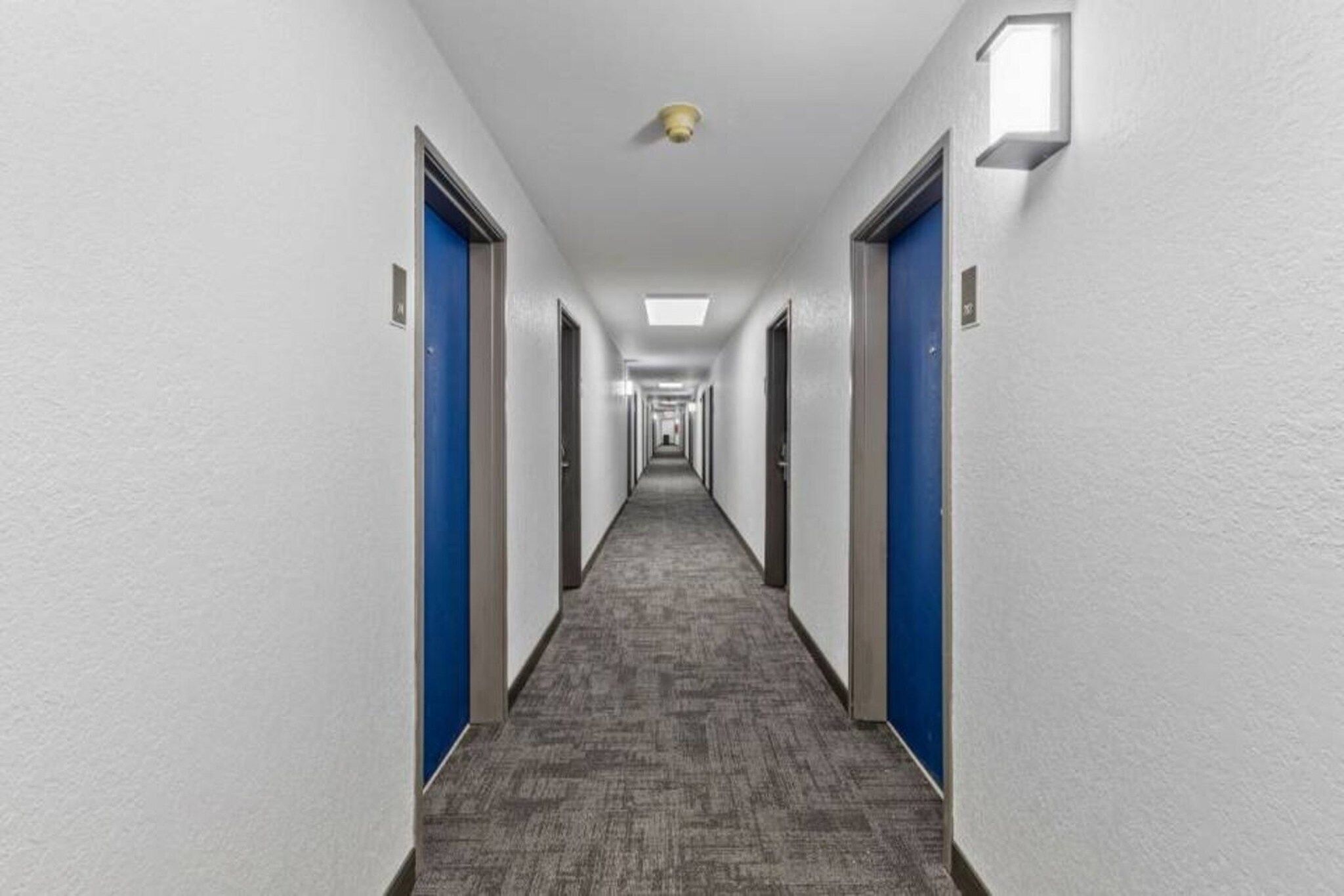 Ein langer, schmaler Hotelkorridor mit Türen an beiden Seiten, Teppichboden und einer Deckenleuchte.