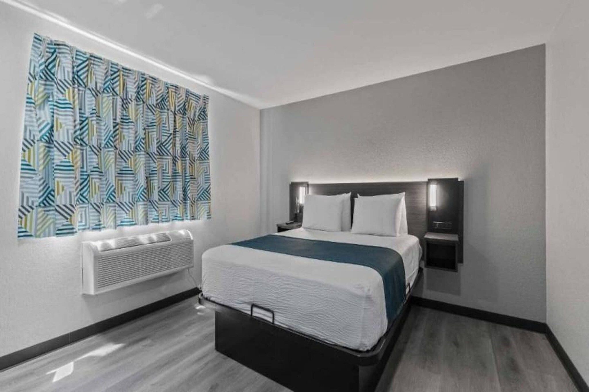 A hotel room with a bed, a wall-mounted heater, and a patterned curtain.