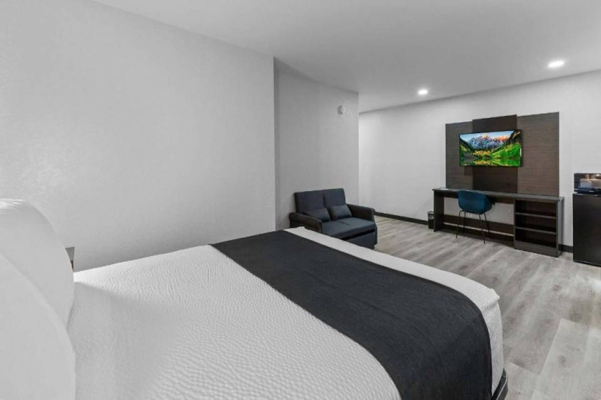 Ein modernes Hotelzimmer mit einem großen Bett, einem Schreibtisch mit Stuhl, einem an der Wand befestigten Fernseher und einer kleinen Küchenzeile.