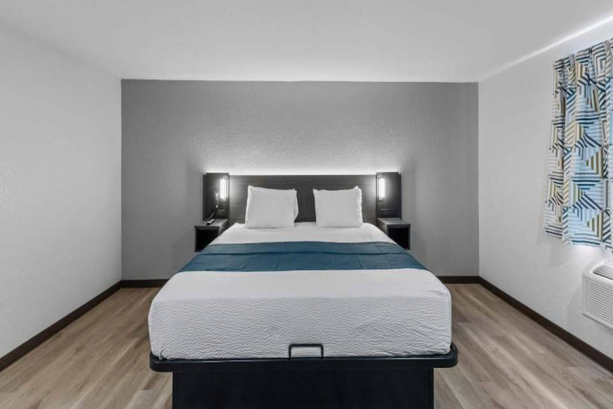 A hotel room with a bed, bedside tables, a headboard with integrated lighting, a wall-mounted air conditioning unit, and a patterned wall decoration.