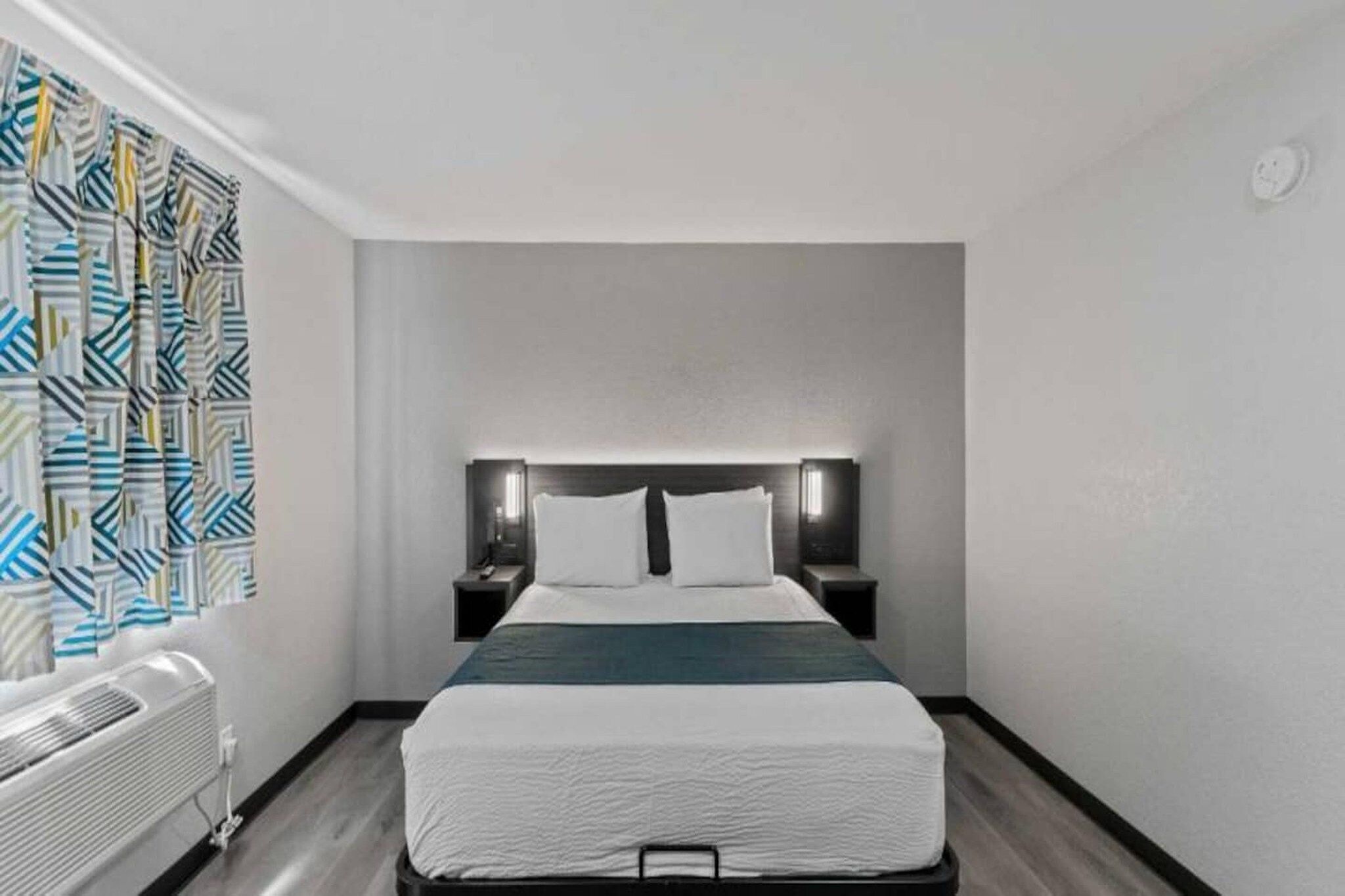 A hotel room with a bed, bedside tables, a wall-mounted heater, and a patterned curtain.