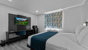 A modern hotel room with a bed, a desk with a chair, a television, and a patterned window treatment.