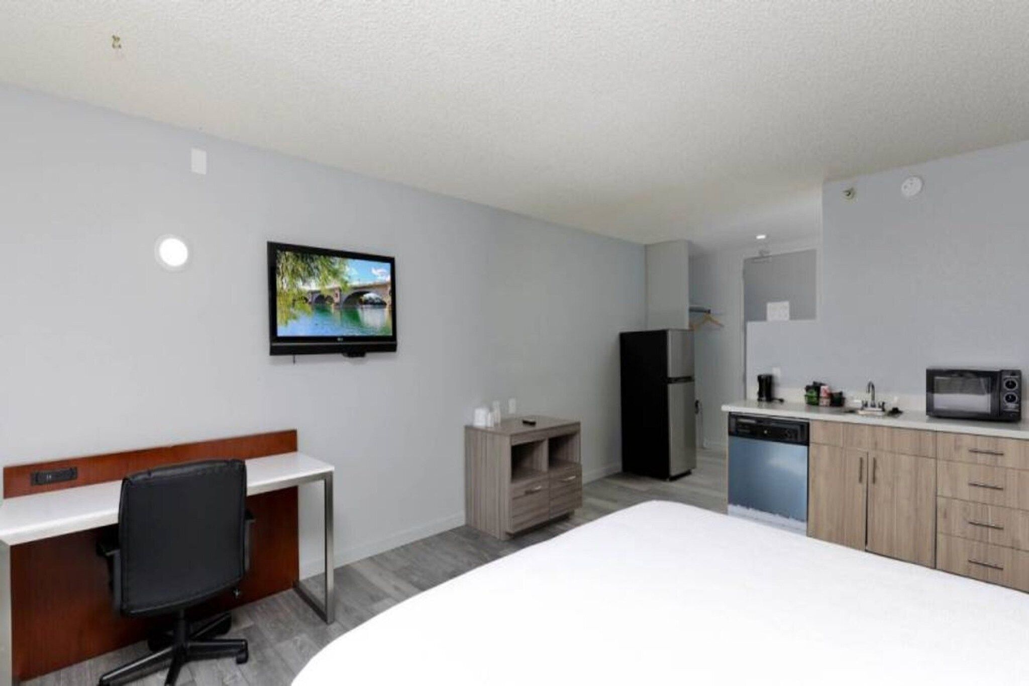 A hotel room with a bed, a desk, a chair, a television, a refrigerator, a microwave, and a small kitchenette.