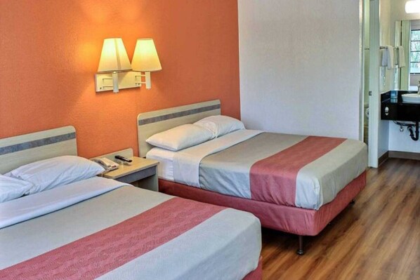 Una camera d'albergo con due letti, un comodino, una lampada e un bagno con lavandino e specchio.
