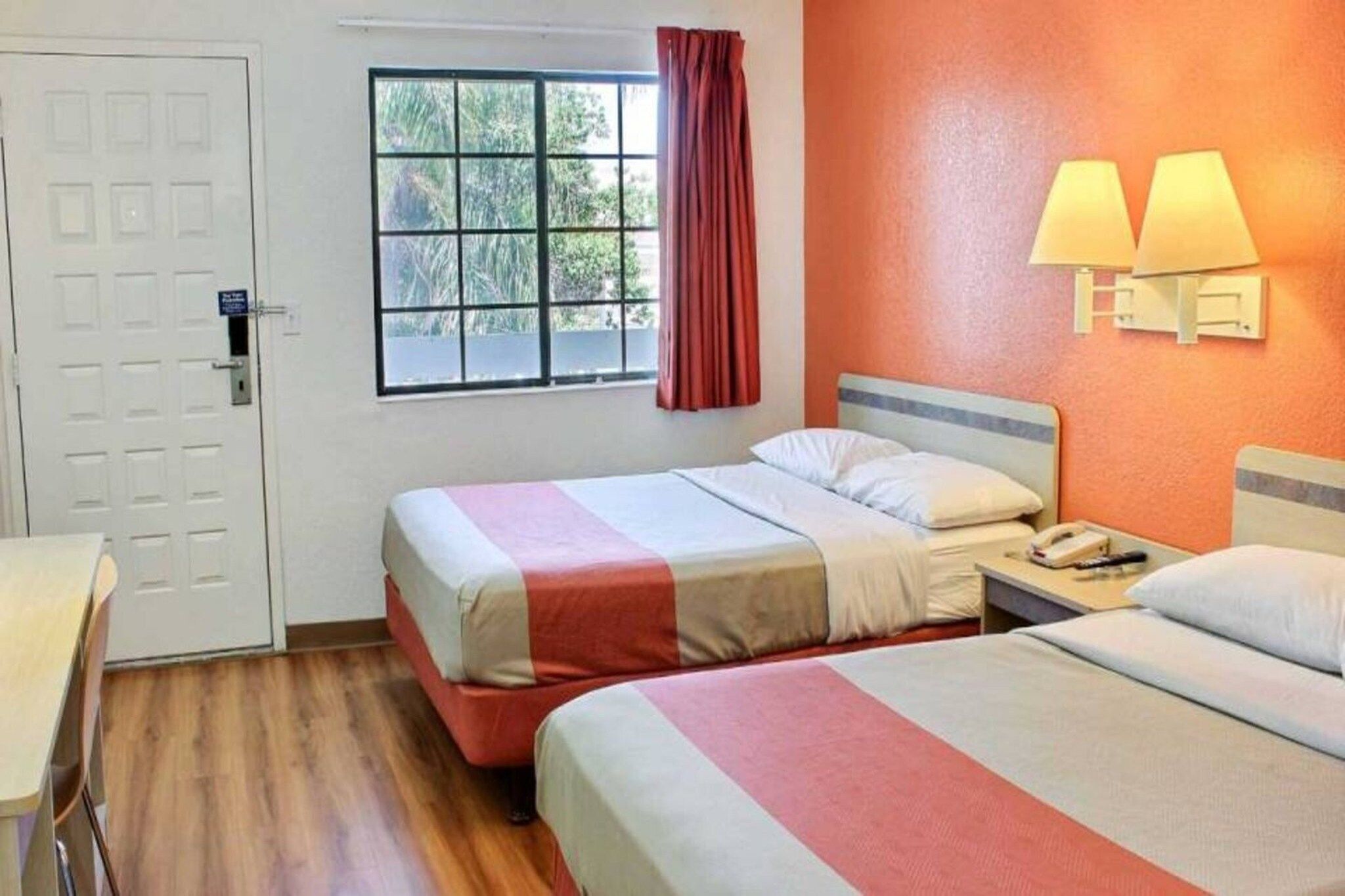 A hotel room with two beds, a window with a view of trees, a door, and a nightstand with a lamp.