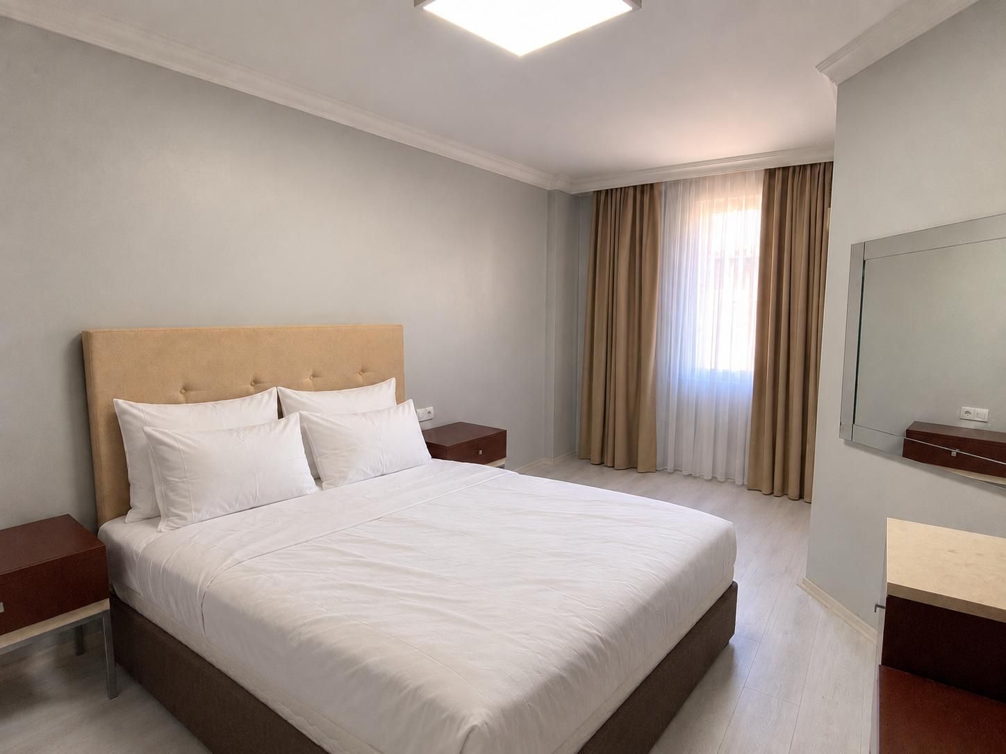 A hotel room with a large bed, a bedside table, a nightstand, a mirror, and a window with curtains.