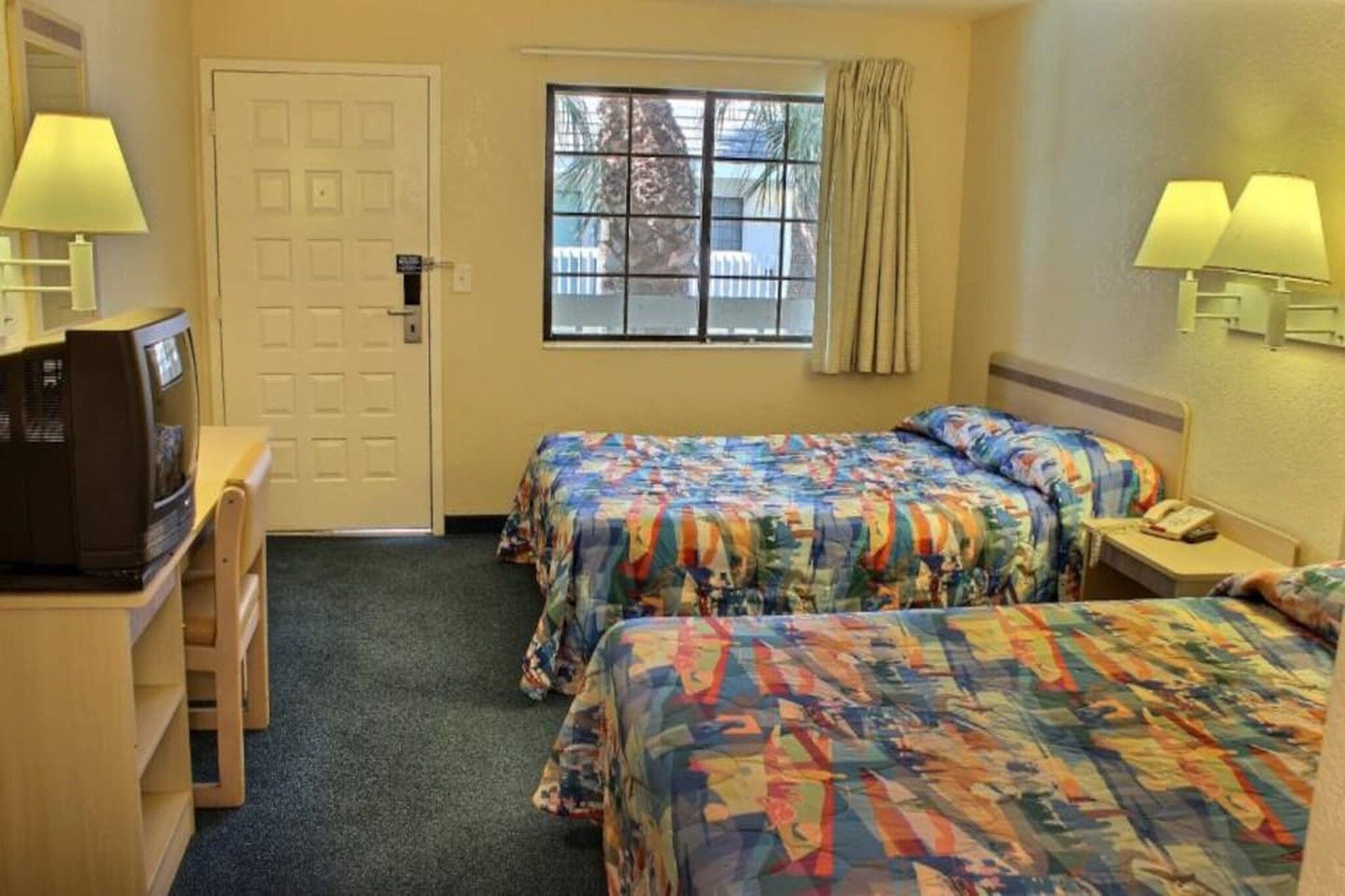 A hotel room with two beds, a desk with a television, a window with curtains, and a door.