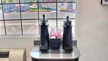 A coffee station with two espresso machines and a coffee grinder, located near a window with a view of a parking lot and buildings.