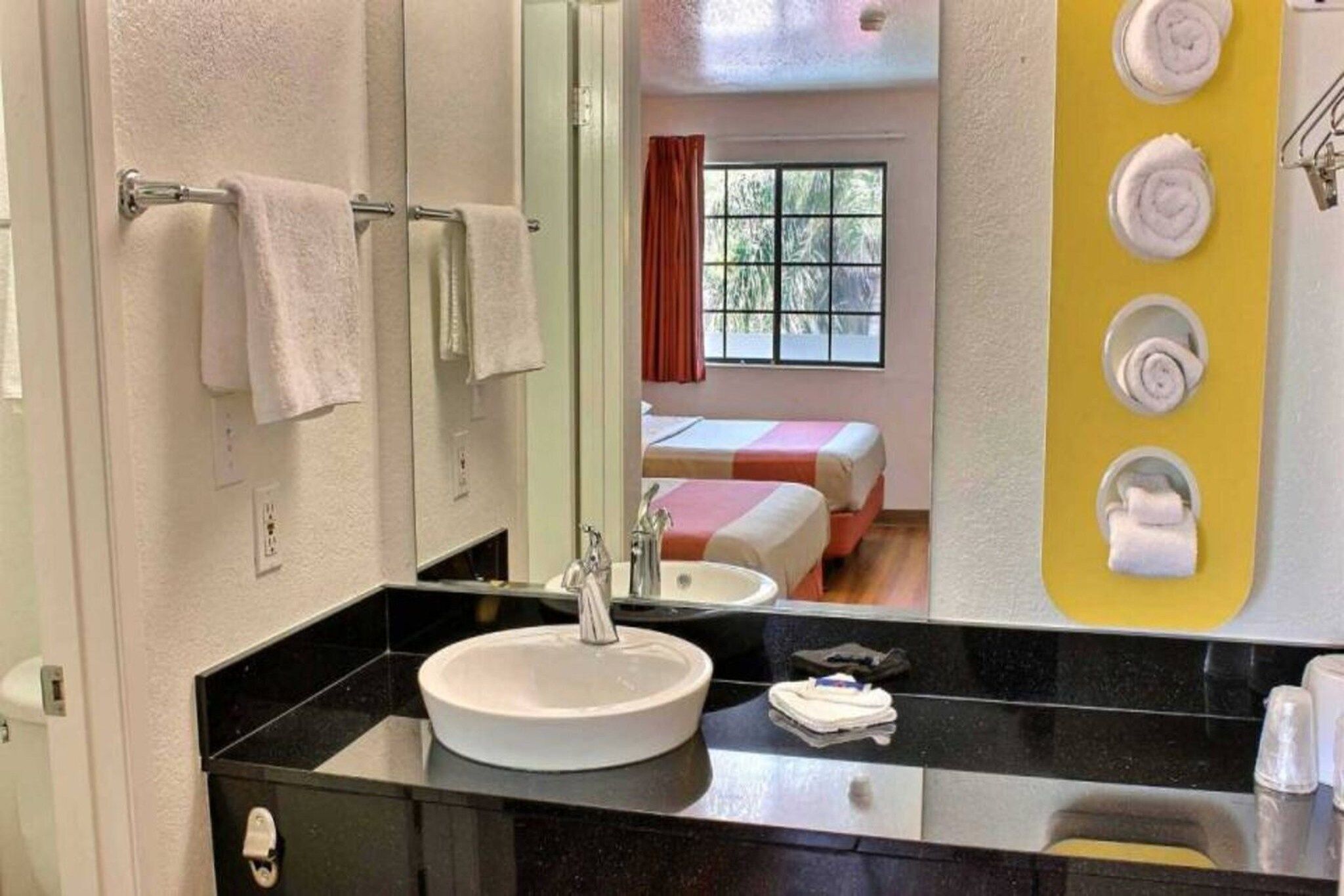 A hotel bathroom with a sink, mirror, towel rack, and a bed visible through the window.