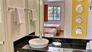 A hotel bathroom with a sink, mirror, towel rack, and a bed visible through the window.