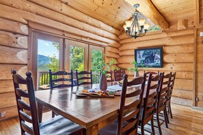 A wooden cabin dining area with a table set for six, surrounded by chairs, a chandelier, and a framed picture on the wall. - Sleeps 24 • 2 Cabins • Hot Tubs + Game Rooms + Mtn Views | Bear Ridge Cabins (Gatlinburg)