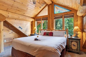 A wooden cabin bedroom with a large bed, two bedside lamps, and a window with a view of trees. - Sleeps 24 • 2 Cabins • Hot Tubs + Game Rooms + Mtn Views | Bear Ridge Cabins (Gatlinburg)