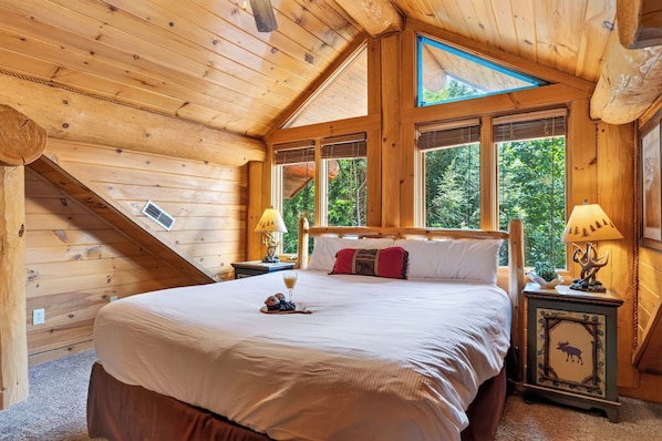 Ein Schlafzimmer einer Holzhütte mit einem großen Bett, zwei Nachttischlampen und einem Fenster mit Blick auf Bäume.