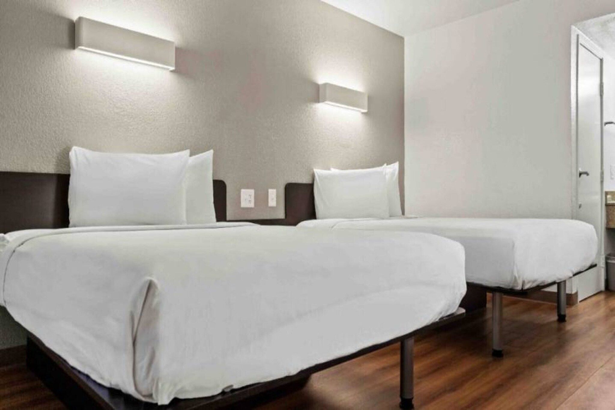 Hotellihuone, jossa on kaksi sänkyä, puulattia ja seinään asennetut valaisimet.