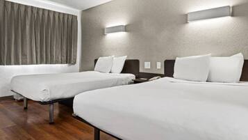 A hotel room with two beds, a window with curtains, a wooden floor, and wall-mounted lighting.