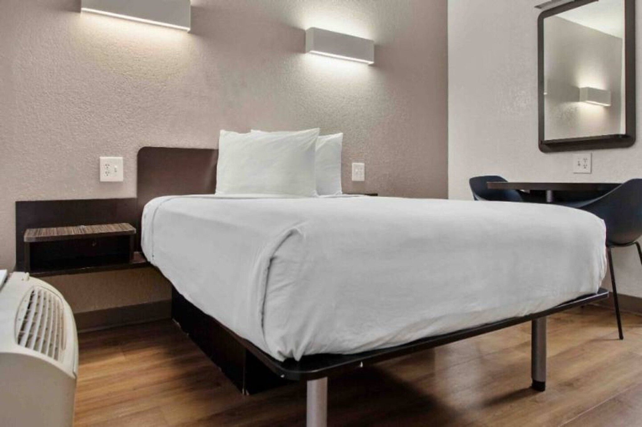 Een hotelkamer met een bed, nachtkastje, stoel en een spiegel aan de muur.