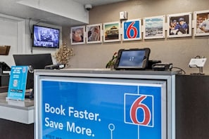 Een receptiebalie van een hotel met een groot scherm waarop een promotieboodschap staat, een televisie en een reeks ingelijste foto's aan de muur.