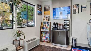 Una habitación con ventana, una planta, un estante en la pared con libros, un refrigerador y un cubo de basura.