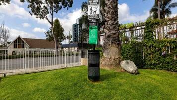 A pet waste station with a sign, a trash bin, a tree, and a fence.