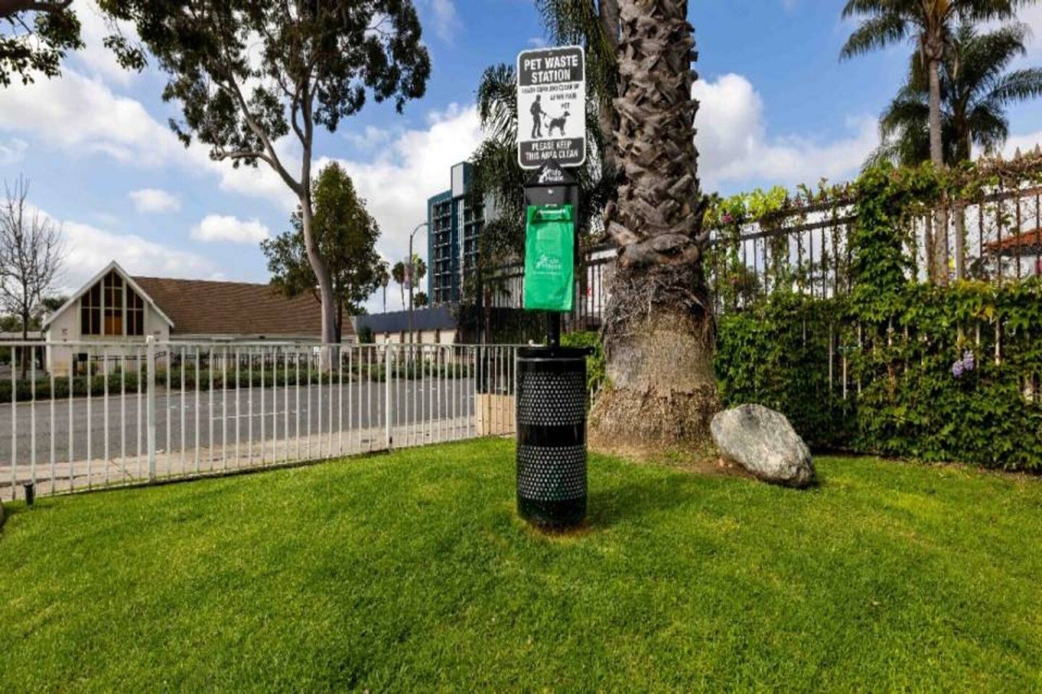 A pet waste station with a sign, a trash bin, a tree, and a fence.