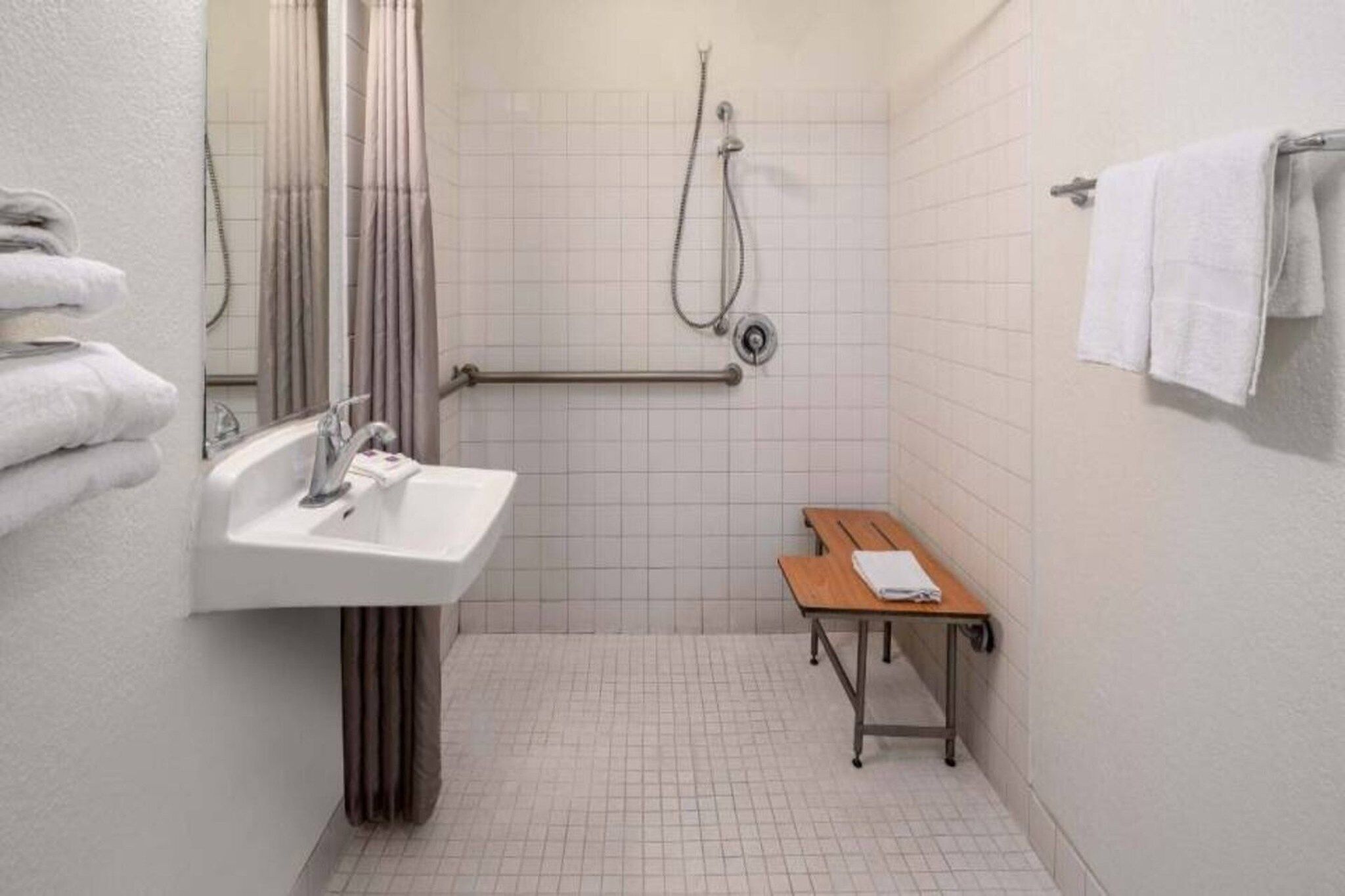 Ein kleines, sauberes Badezimmer mit Waschbecken, Spiegel, Dusche und einer hölzernen Sitzbank.