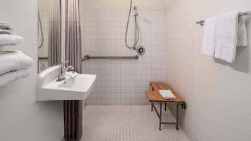 A small, clean bathroom with a sink, mirror, shower, and a wooden bench.
