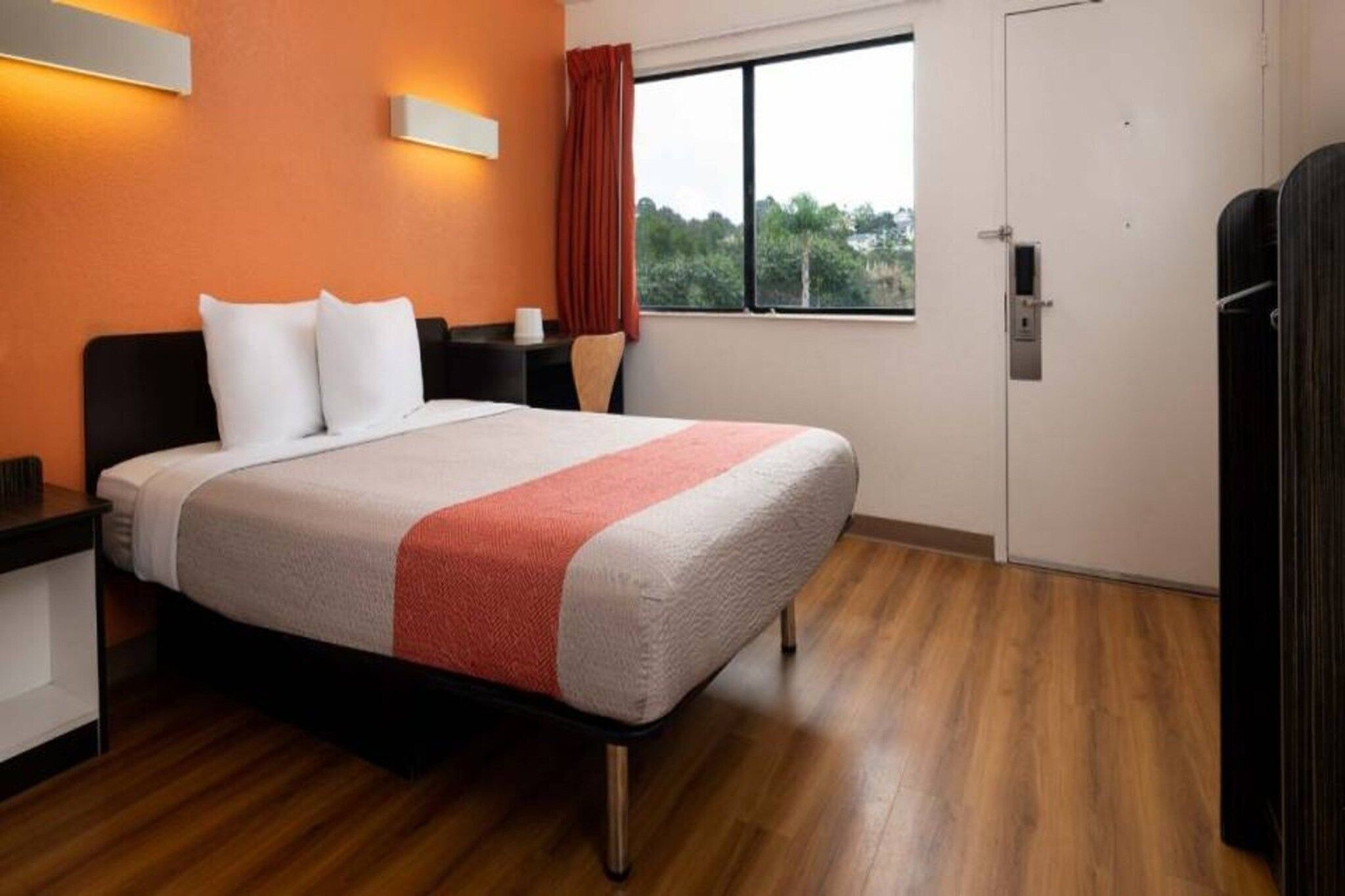 A hotel room with a bed, a nightstand, a window with curtains, and a door.