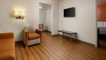 A hotel room with a wooden floor, a sofa, a chair, a small table, and a TV mounted on the wall.